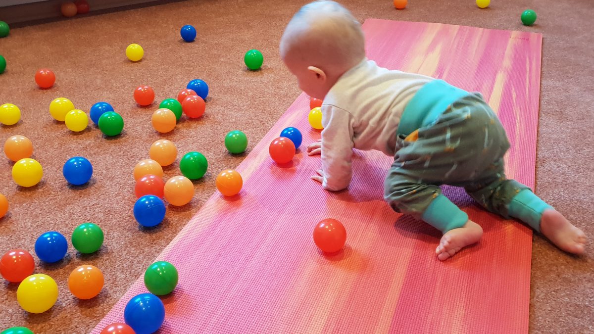 Ein Baby krabbelt über eine Yogamatte, bunte Bälle sind auf dem Boden verteilt