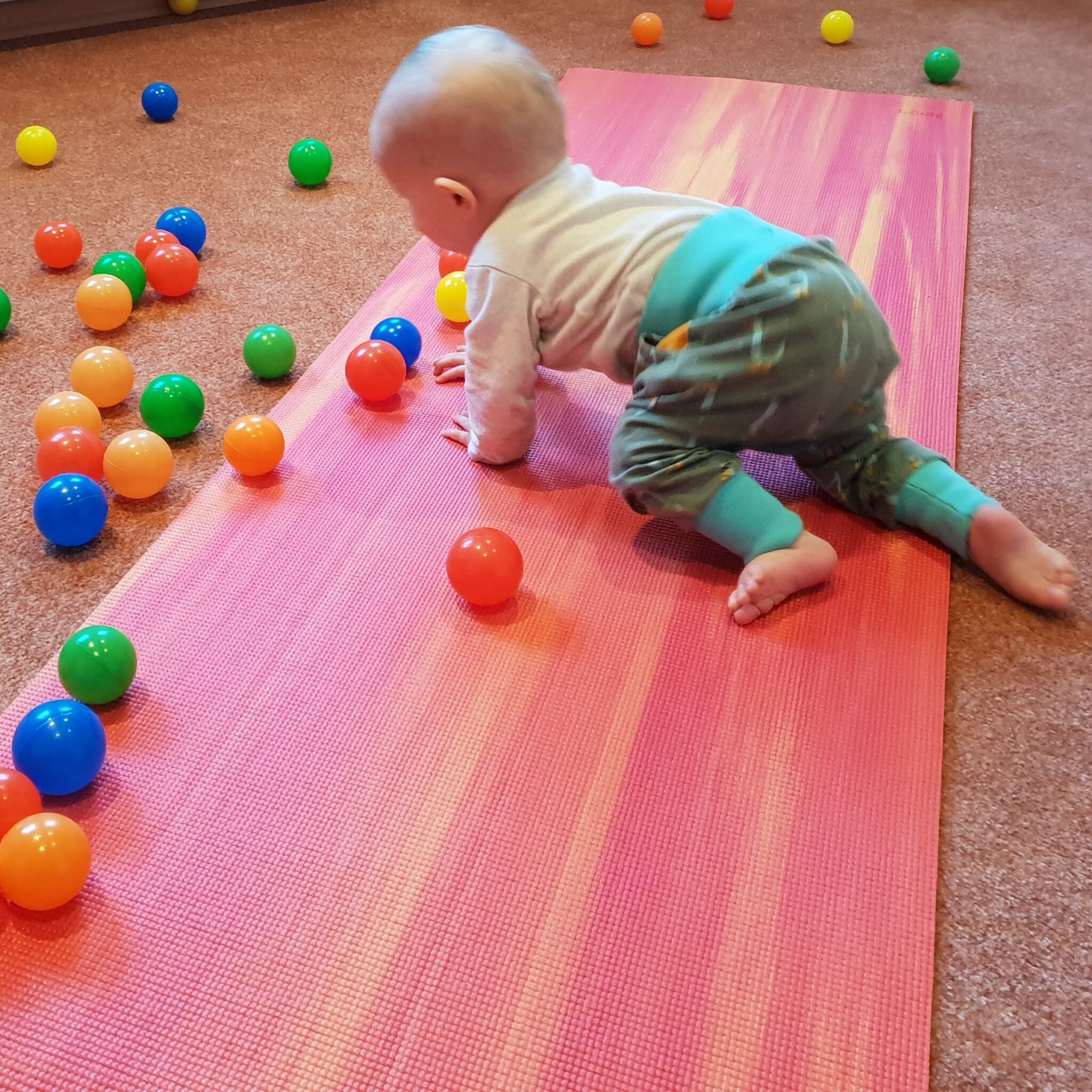 Ein Baby krabbelt über eine Yogamatte, bunte Bälle sind auf dem Boden verteilt