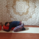 Marion übt Yin Yoga und liegt in der position der Frosch. Das Gesäß ist auf den Fersen, die Knie sind leicht nach außen abgelegt. Der Oberkörper ruht vorn auf einer Yogarolle.