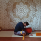 Marion übt Yin Yoga und übt die Position vorgebeugter Schmetterling. Die Fußsohlen sind aneinandergelegt, ein Yogagurt ist um die Knie gelegt und stützt den Bereich. Die Stirn ist abgesenkt und ruht auf zwei Kissen.