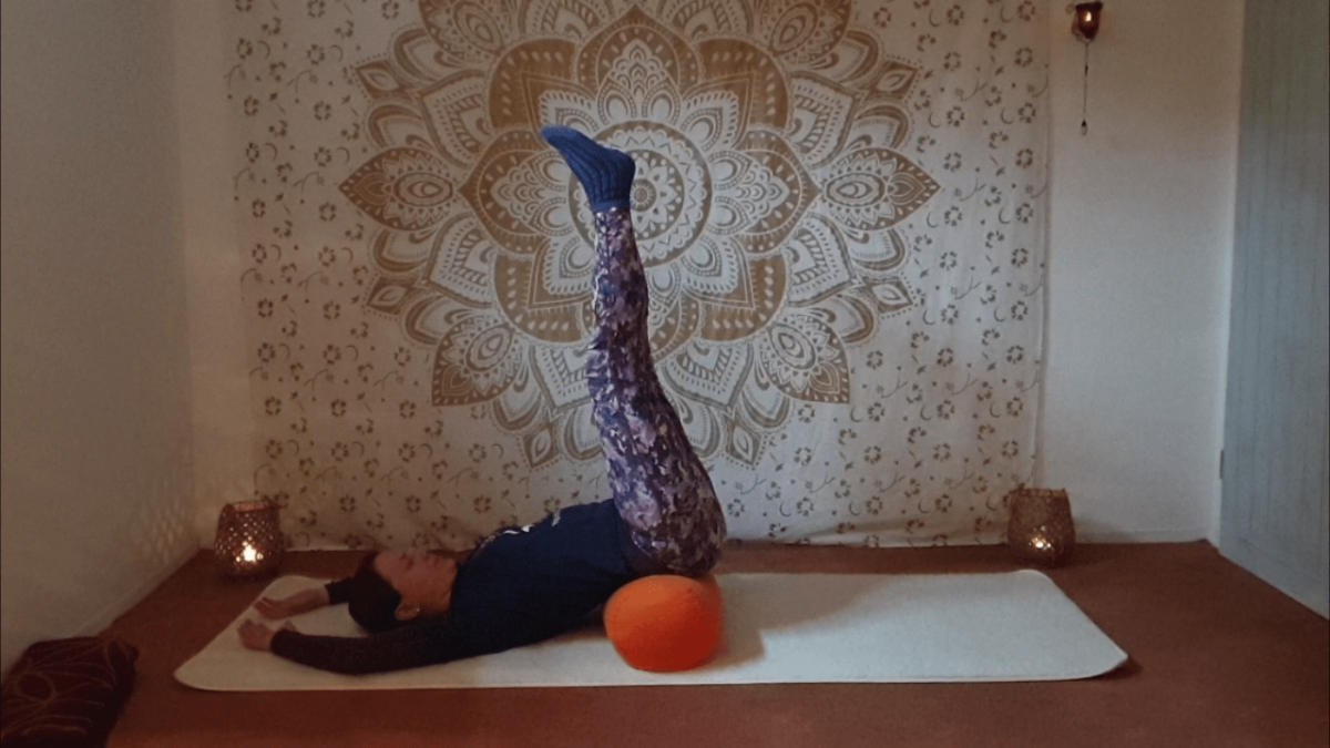 Marion übt Yin Yoga und die Position Seegras. Sie liegt auf dem Rücken, die Arme über dem Kopf abgelegt. Die Beine sind locker zur Decke gestreckt, das Becken liegt erhöht auf einer Yogarolle.