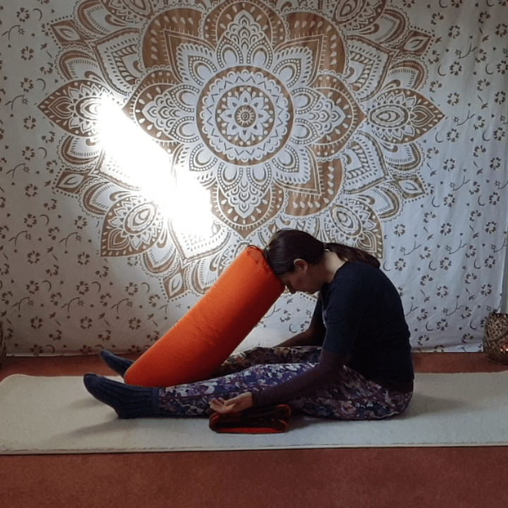 Marion übt Yin Yoga und die Position Raupe. Sie sitzt auf der Matte, die Beine sind lang ausgestreckt und eine Decke ist unter den Knien. Der Oberkörper ist abgesenkt und die Stirn ruht auf einem Yogakissen.
