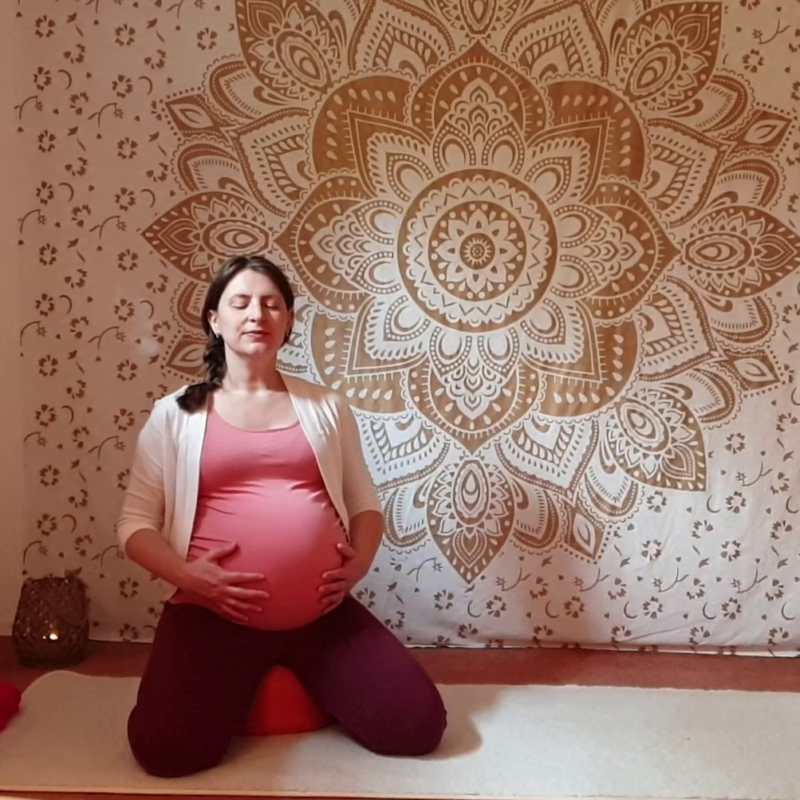 Marion sitzt hochschwanger im Fersensitz auf der Yogamatte, die Hände am Bauch, um den Atem zu spüren. Die Augen sind geschlossen.